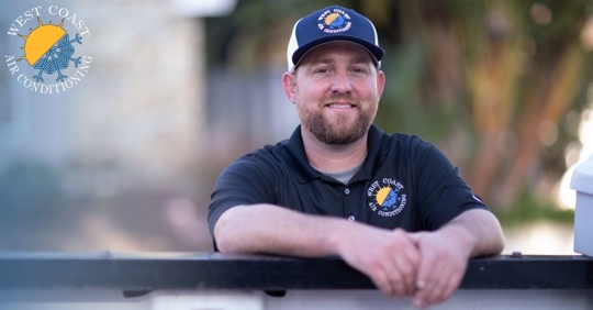 A man in West Coast Air Conditioning hat and shirt leaning on the bed of a pickup truck smiling